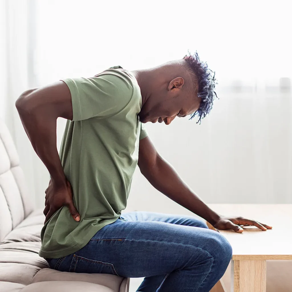 A person in a green shirt and blue jeans sits on a sofa, holding their lower back in pain, leaning forward with a pained expression, suggesting discomfort.